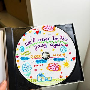 May include: A white CD with colorful drawings on it. The text on the CD says "We'll never be this young again 2000 MIX".