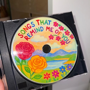 May include: A colorful CD with a hand-drawn illustration of a landscape with flowers, mountains, and a lake. The text "Songs That Remind Me Of You" is written in rainbow colors around the edge of the CD.