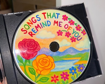 May include: A colorful CD with a hand-drawn illustration of a landscape with flowers, mountains, and a lake. The text "Songs That Remind Me Of You" is written in rainbow colors around the edge of the CD.
