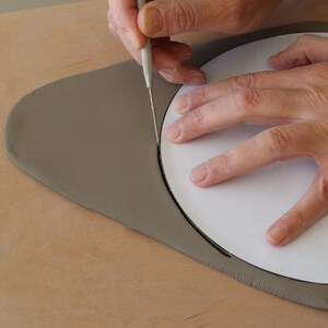 May include: A person using a tool to cut a circle into a piece of gray clay.