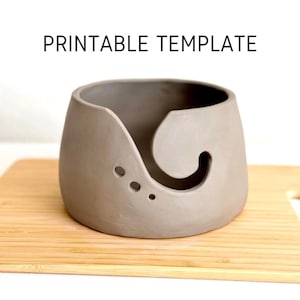 May include: A gray ceramic yarn bowl with a curved opening and three small holes. The bowl is sitting on a wooden cutting board. The text "PRINTABLE TEMPLATE" is above the bowl.