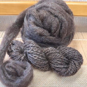 May include: Close-up of brown yarn in various stages: a large, fluffy ball, a skein, and a small, tightly wound ball. The yarn has a soft, textured appearance, suggesting it's made from natural fibers. The background shows a wooden loom.