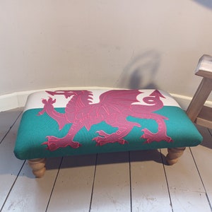 May include: A green and white footstool with a red dragon embroidered on the top. The footstool has four wooden legs.