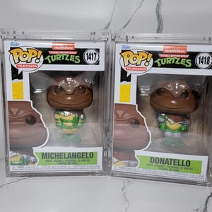 Op de afbeelding: Twee Teenage Mutant Ninja Turtles Pop! Vinyl figuren in beschermende koffers. De figuren zijn Michelangelo en Donatello. Michelangelo draagt een groene bandana en Donatello een paarse bandana. Beide figuren houden hun kenmerkende wapens vast.