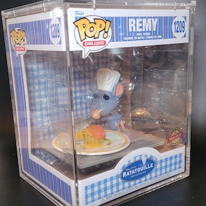 May include: A Funko Pop Deluxe vinyl figure of Remy from the movie Ratatouille. The figure is in a clear display case with a blue and white checkered pattern. Remy is depicted with a chef's hat, preparing food on a plate.