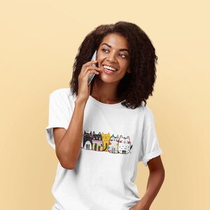 Puede incluir: Camiseta blanca con una fila de gatos de dibujos animados coloridos. Cada gato tiene un diseño único, con patrones y colores como negro, amarillo y azul. La camiseta está hecha de un material suave.