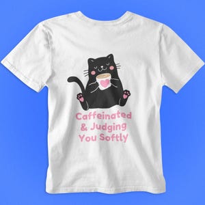 Op de afbeelding: Wit t-shirt met een zwarte cartoonkat die een koffiekopje met een roze hart vasthoudt. De tekst "Caffeinated & Judging You Softly" is in roze onder de kattenillustratie gedrukt.