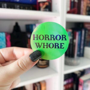 以下が含まれることがあります： 黒文字で「HORROR WHORE」と書かれた緑色の虹色のステッカーです。
