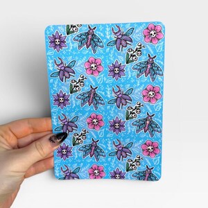 May include: A rectangular sticker sheet with a bright blue background featuring a repeating pattern of purple moths, blue beetles, pink flowers with skull centers, and small white floral sprigs, all outlined in bold black ink lines.