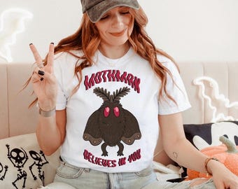May include: A white short-sleeve t-shirt featuring a graphic of a brown moth with red eyes, surrounded by the red text "MOTHMAN BELIEVES IN YOU". The shirt is paired with a charcoal gray baseball cap with the text "STAY SPOOKY" and a skeleton hand.
