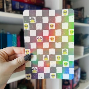 Peut inclure: Un autocollant holographique avec un motif à carreaux et des visages d'extraterrestres. L'autocollant a une finition arc-en-ciel irisée.