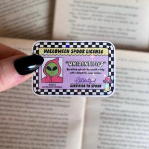 May include: A holographic sticker featuring a cartoon alien wearing a pink hoodie. The sticker is a Halloween Spook License and reads "UNIDENTIFIED" with the text "Certified out-of-this-world entity with a knack for crop circles." The sticker is signed "Undentified" and reads "CERTIFIED TO SPOOK".