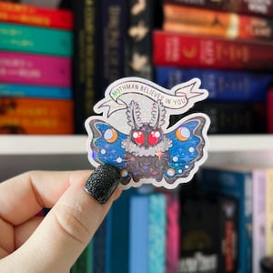 May include: A holographic sticker featuring a mothman with iridescent blue wings and red eyes. The sticker has the text "Mothman Believes in You" on a banner above the mothman.