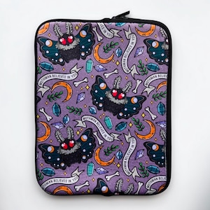 May include: Purple laptop sleeve with a repeating pattern of moths, crescent moons, bones, and crystals. The text "Mothman Believes in You" is repeated throughout the pattern.