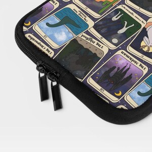 Puede incluir: Una funda negra con cremallera que presenta una cuadr&iacute;cula de cartas estilo tarot con ilustraciones de cr&iacute;ptidos. Las cartas visibles incluyen The Loch Ness Monster, The Bigfoot y The Mothman, con ilustraciones, n&uacute;meros romanos y texto.