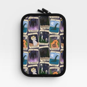 Puede incluir: Un estuche rectangular negro con cremallera que presenta un patr&oacute;n repetitivo de cartas estilo tarot. Cada carta representa un cr&iacute;ptido diferente, incluyendo Mothman, Bigfoot, el monstruo del Lago Ness, el Jackalope y el Chupacabra, sobre un fondo morado.