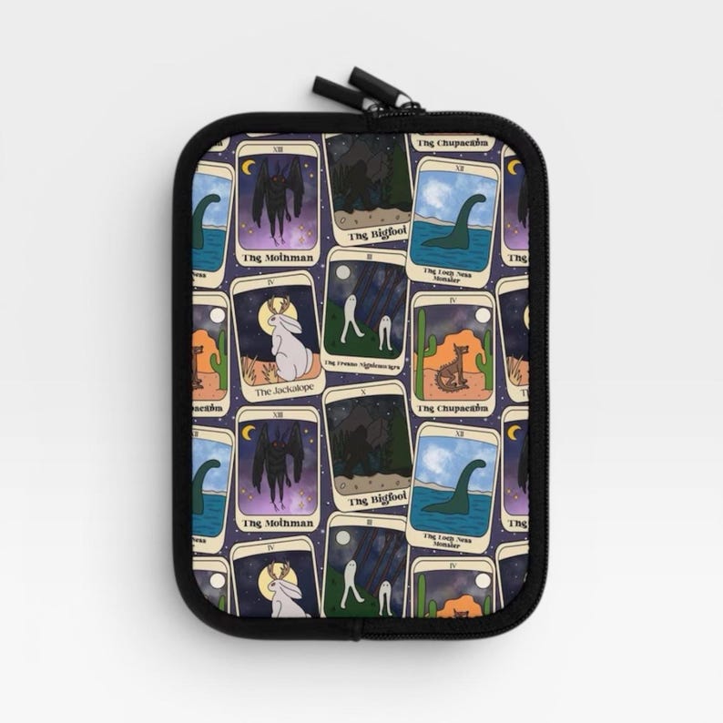 Puede incluir: Un estuche rectangular negro con cremallera que presenta un patr&oacute;n repetitivo de cartas estilo tarot. Cada carta representa un cr&iacute;ptido diferente, incluyendo Mothman, Bigfoot, el monstruo del Lago Ness, el Jackalope y el Chupacabra, sobre un fondo morado.