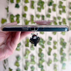 May include: A clear phone case with a black and white cartoon character charm attached to the headphone jack. The character has red eyes and black wings.