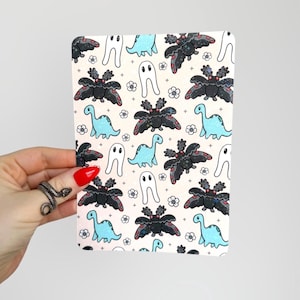 Peut inclure: Un bloc-notes rectangulaire avec un motif répété de dinosaures, de papillons de nuit, de fantômes et de fleurs de dessins animés en noir, bleu et blanc sur un fond rose clair. Le bloc-notes est tenu par une main avec un ongle peint en rouge et une bague serpent.