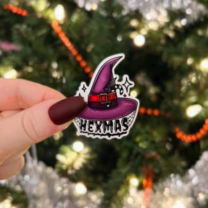 May include: A purple witch hat with a red band and a candy cane. The hat is on a white sticker with the word "Hexmas" written in black.