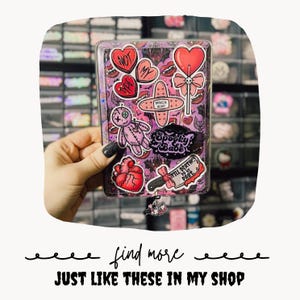 May include: A rectangular clear plastic card holder decorated with various stickers including a pink voodoo doll, a red anatomical heart, a lollipop, a bandage labeled broken heart, and a knife with the text Till Death Do Us Part.