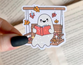 May include: A white ghost with a happy face sits on a wooden swing reading a red book.  Fall leaves are scattered around the ghost and swing.