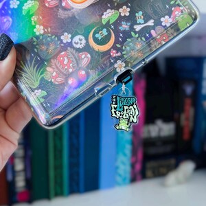 May include: A clear phone case with a colorful, whimsical design featuring mushrooms, flowers, and a crescent moon. A charm with a green frog and the text "The Legend Froggy" is attached to the case.