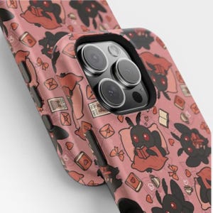 May include: A phone case with a dusty pink background featuring a repeating pattern of black moth-like creatures with red eyes, some reading books, alongside small red hearts, envelopes, bows, and coffee cups. The case has a black camera lens cutout.