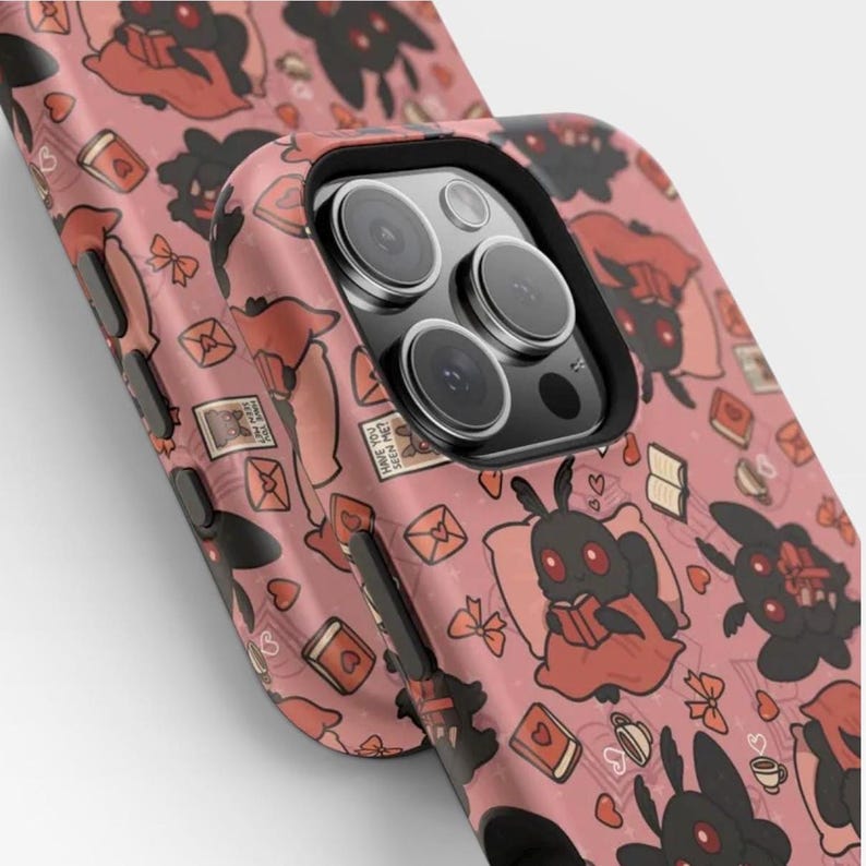 May include: A phone case with a dusty pink background featuring a repeating pattern of black moth-like creatures with red eyes, some reading books, alongside small red hearts, envelopes, bows, and coffee cups. The case has a black camera lens cutout.