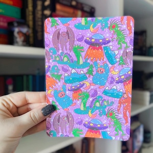 Peut inclure: Un fond violet avec un motif répété de monstres, d'extraterrestres et de montagnes colorés. Les monstres sont bleus, verts et orange. Les extraterrestres sont des soucoupes volantes avec des accents orange et jaunes. Les montagnes sont bleues et vertes. Le fond est recouvert de paillettes.