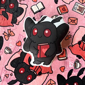 May include: A plush toy shaped like a black moth with red eyes holding a red book with a heart on it. The moth is sitting on a pink and white patterned background with other moths and books.