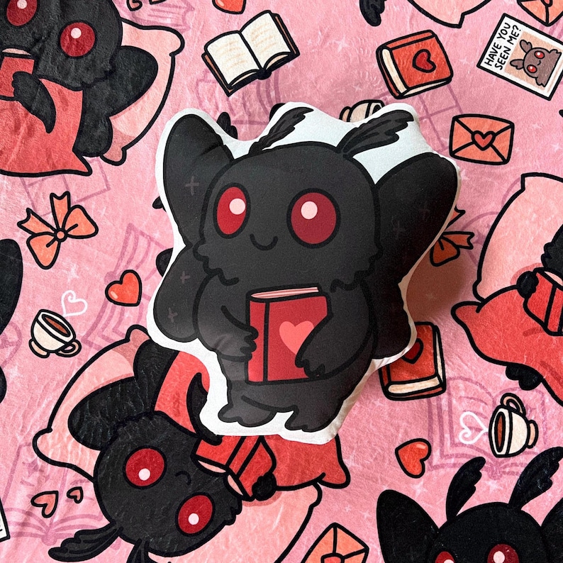 May include: A plush toy shaped like a black moth with red eyes holding a red book with a heart on it. The moth is sitting on a pink and white patterned background with other moths and books.