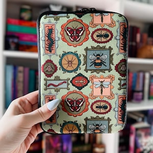 May include: A zippered book sleeve with a light green background and a repeating pattern of framed insects and butterflies in various colors. The sleeve is held by a person.