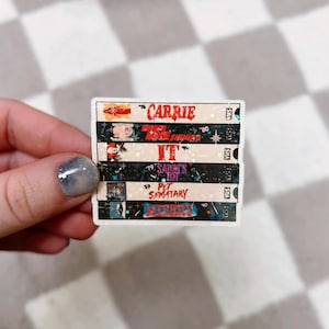 May include: A sticker featuring a stack of VHS tapes with titles including "Carrie," "The Shining," "IT," "Salem's Lot," "Pet Sematary," and "Misery." The tapes have a vintage aesthetic with colorful graphics and text.