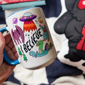 May include: White ceramic mug with a blue rim. The mug features a colorful illustration of a monster, a UFO, a creature from the black lagoon, and the words "I Believe".