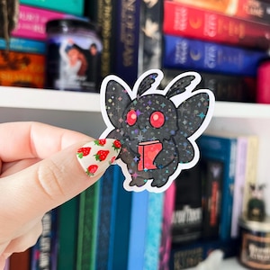 May include: A black and red holographic sticker of a moth reading a book. The moth has sparkly wings and red eyes.