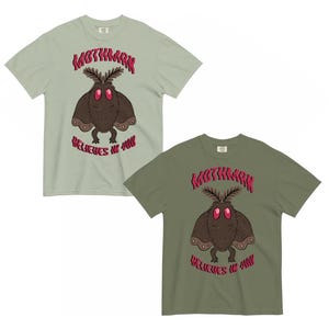 K&ouml;nnte beinhalten: Zwei kurz&auml;rmelige T-Shirts in hellem Salbeigr&uuml;n und Olivgr&uuml;n, jeweils mit einer zentralen Grafik einer braunen, mottenartigen Kreatur mit gro&szlig;en roten Augen, dar&uuml;ber der Text "MOTHMAN" und der Schriftzug "BELIEVES IN YOU" in roter Farbe.