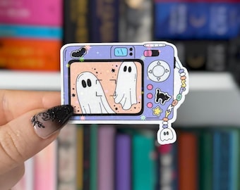 Puede incluir: Una pegatina de cámara digital de color morado claro con una pantalla que muestra dos fantasmas blancos sobre un fondo melocotón. El cuerpo de la cámara incluye un murciélago negro, un gato negro, una estrella amarilla y una correa de cuentas.