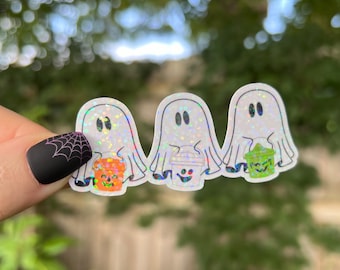 May include: Three white ghost stickers with holographic glitter, each holding a different container: a jack-o'-lantern, a coffee cup, and a green bucket. The stickers have black oval eyes and are set against a blurred green background.