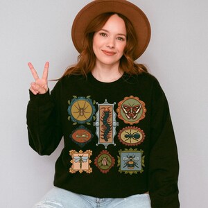 May include: A black long-sleeve sweatshirt featuring a graphic print of nine various insects and arachnids, each enclosed in its own decorative, ornate frame. The creatures include a spider, centipede, butterfly, mantis, ant, dragonfly, and bee.