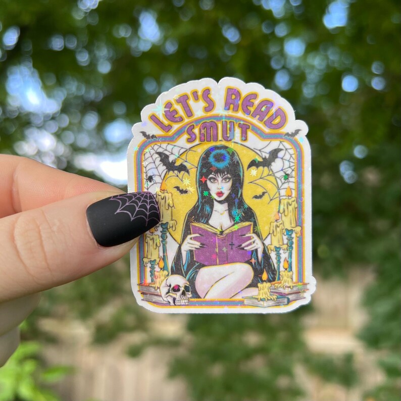 May include: A sticker with the text "LET'S READ SMUT" above an illustration of a woman reading a purple book. The image has a spooky theme with bats, candles, and a skull. The sticker has a white border.