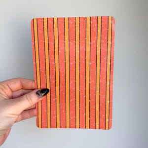 May include: A rectangular card featuring a vertical striped pattern in alternating shades of coral orange and bright orange. The design is accented with thin black vertical lines and a subtle, scattered pattern of small white four-pointed stars.