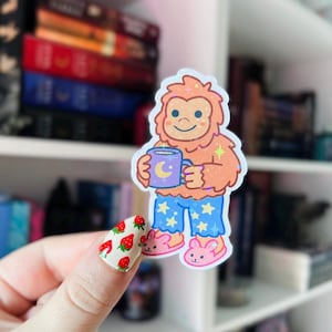 Op de afbeelding: Een sticker met een cartoonfiguur dat een pyjama draagt en een mok vasthoudt met een halve maan erop. Het figuur heeft oranje vacht en een blauwe pyjama met witte sterren. De sticker is op een witte achtergrond.