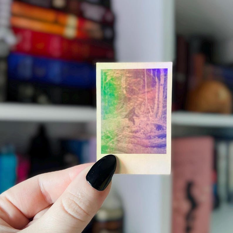 May include: A small, square sticker with a holographic image of a forest scene. The sticker has a rainbow gradient effect, with colors blending from green to purple. The image is held by a hand with a black nail.