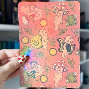 May include: Pink background with a repeating pattern of fairies, mushrooms, frogs, and strawberries. The fairies are nude and have wings. The mushrooms are red and white. The frogs are green. The strawberries are red.