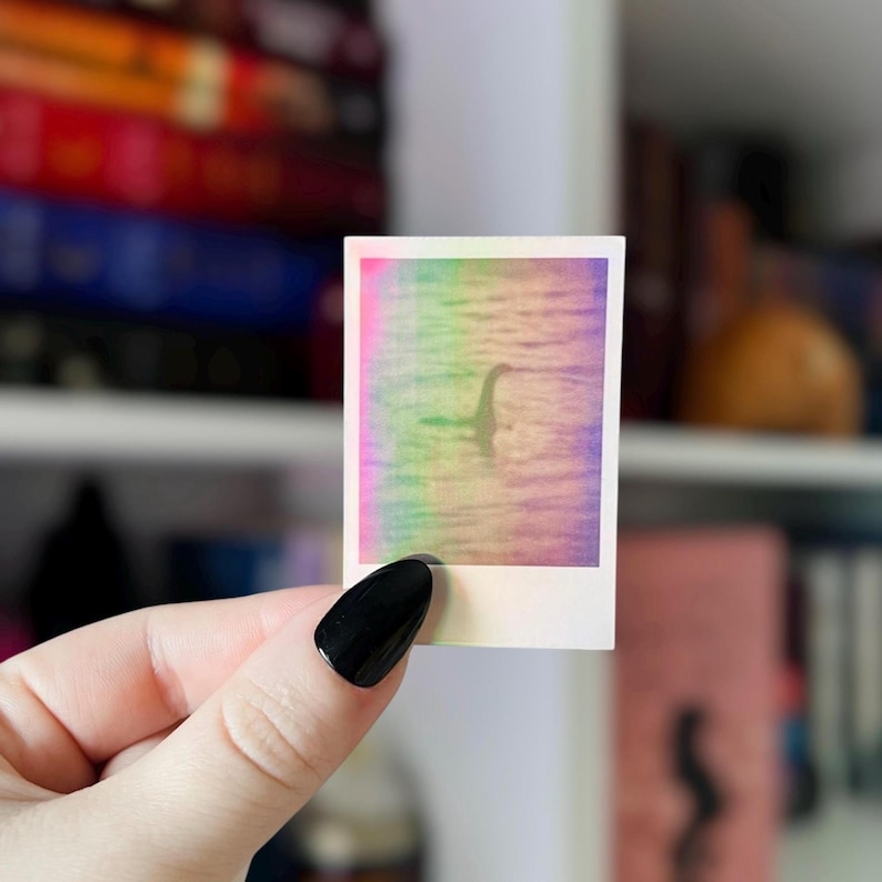May include: A small, square sticker featuring a holographic image of the Loch Ness Monster. The image has a rainbow gradient background. The sticker is held by a hand with a black painted nail.