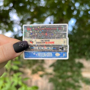 May include: A sticker of a stack of VHS tapes with the titles "The Craft", "Poltergeist", "The Texas Chainsaw Massacre", "The Exorcist", and "Hellraiser".