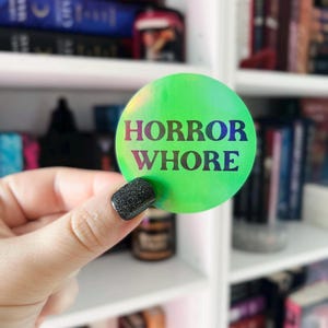 以下が含まれることがあります： 黒い文字で「HORROR WHORE」と書かれた緑色の虹色のステッカーです。