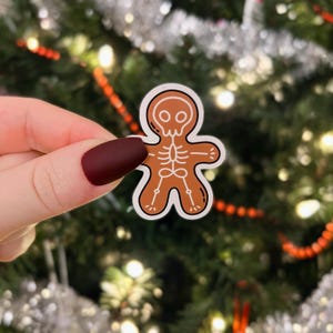 May include: A gingerbread man sticker with a skeleton design. The sticker is brown with white outlines and is held in a hand against a blurry background of a Christmas tree.