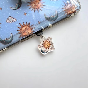 May include: A clear phone case with a celestial design featuring suns, moons, clouds, and stars. A matching charm with a sun, moon, and cloud design is attached to the case. The background of the case is light blue.
